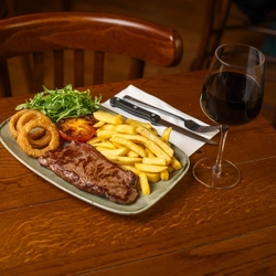 Steak & Chips served on a plate with salad, tomato, and onion rings. The plate sits on a wooden restaurant table along with cutlery, a napkin, and a glass of red wine.