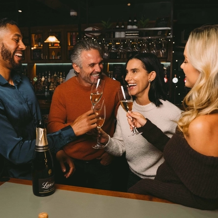 Four people standing at a table in a bar, laughing together and raising glasses of champagne. A bottle of Lanson champagne stands on the table, and a Christmas tree decorated in red and gold stands beside them.