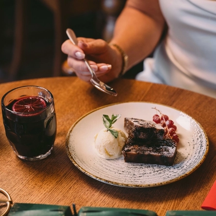 A close up view of two slices of Christmas pudding served on a plate with berries and a scoop of ice cream. The plate sits on a wooden restaurant table along with two green Christmas crackers and a mug of mulled wine; and a person at the table is holding a spoon above the plate.