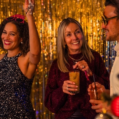 Three people stand together in front of gold and silver metallic party backdrops during a festive New Year’s celebration. One person wears gold novelty party glasses and holds a drink, while the others hold tall glasses with straws. A decorated Christmas tree with red and gold baubles, candy cane ornaments and warm fairy lights is visible in the foreground. The scene is illuminated with warm decorative lighting, creating a festive holiday party setting.
