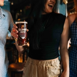 A close up view of two people standing inside a pub; one of them is holding a pint of Tennent's in a branded glass, and the other is holding a pint of Belhaven Best in a branded glass.