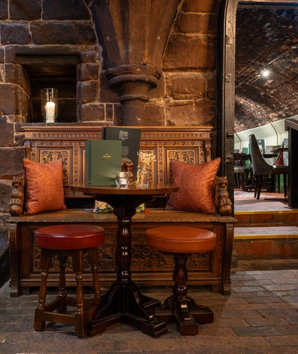 An image focusing on one of the tables within the interior restaurant seating area with ornate wooden carved bench seating, stone walls and brick flooring at The Watergates Bar.