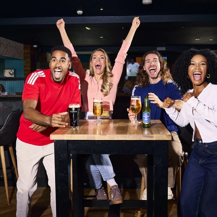 An image showing 4 friends at a table enjoying drinks and celebrating while watching on a football match within the interior seating area at a Flaming Grill venue.