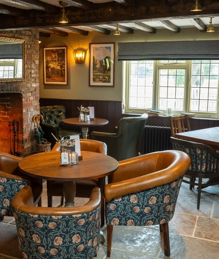 The interior restaurant seating area at The Green Man in Great Offley, with a large brick woodburning fireplace, upholstered chairs, stone floor, and wooden beams on the ceiling.