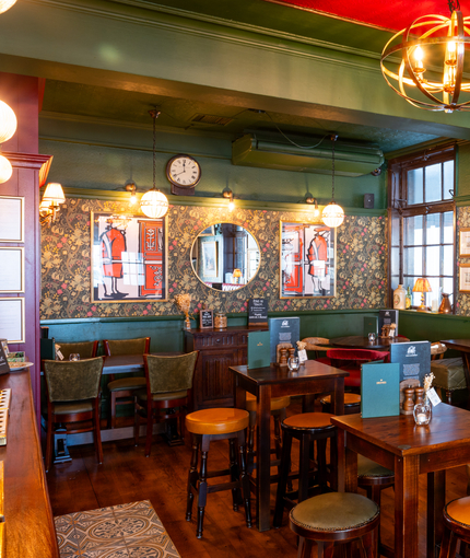 The interior restaurant seating area at the Two Chairmen in St James's, with upholstered chairs and bar stool seating, framed artwork on the floral wallpapered walls, and a fireplace.