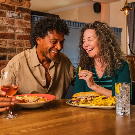 Two people seated at a wooden table in a pub, enjoying lunch and drinks. One plate holds a sandwich with chips, while another contains a salad with sliced vegetables. A glass of rosé wine and a clear drink with ice and lemon are on the table. Exposed brickwork, blue panelled dividers, leather seating and warm pendant lighting create a cosy pub interior, with a window overlooking greenery in the background.