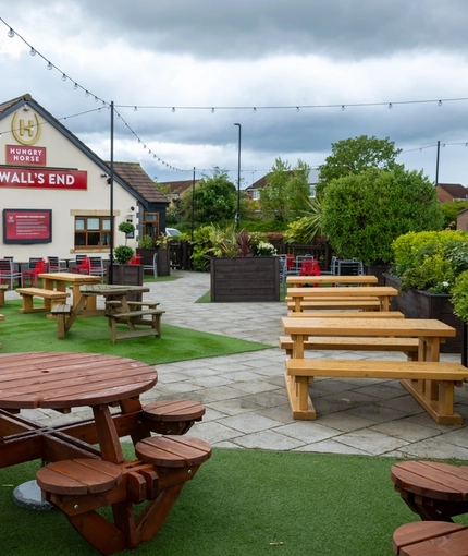 The exterior facade, signage, and beer garden seating area at The Wall's End in Wallsend, with wooden picnic tables, string lights overhead, and a TV on the wall.