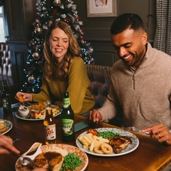 Person in a mustard jumper eating a steak meal with chips, peas and onion rings at a pub table, with a decorated Christmas tree and festive lights in the background. A bottle of Heineken stands beside the plate, while other diners and food are visible in the foreground.