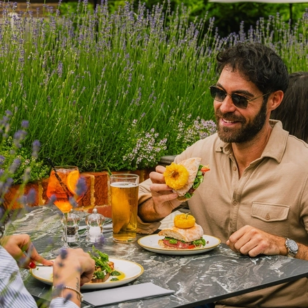 An image of two people enjoying two dishes from the lunch menu and drinks within the exterior beer garden seating area at a Crafted venue.