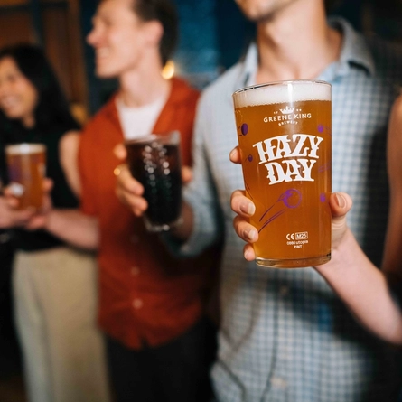 A close up view of a person standing inside a pub and holding a branded glass of Hazy Day. Other people are standing in the background, also holding drinks.