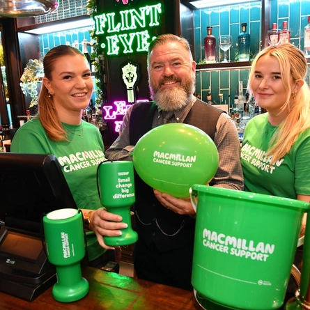 A picture of three Greene King staff members behind a bar with various Macmillan fundraising merchandise.