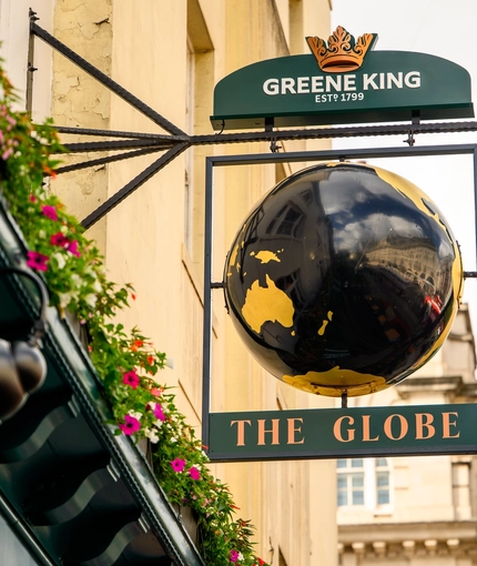 A close up view of the pub sign hanging on the exterior wall of The Globe in Marylebone. The sign features a three-dimensional, black and gold globe within a dark green metal frame below the Greene King logo, and above the words "The Globe" in gold letters.