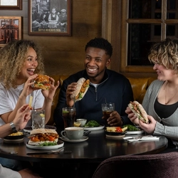 A group of four people sit around a round wooden table in a restaurant. Each person holds a sandwich or burger. The table is set with plates of food including fries topped with melted cheese and garnishes, small bowls of salad or vegetables, and drinks including glasses of dark soda with ice and slices of lemon, as well as mugs and cups. 