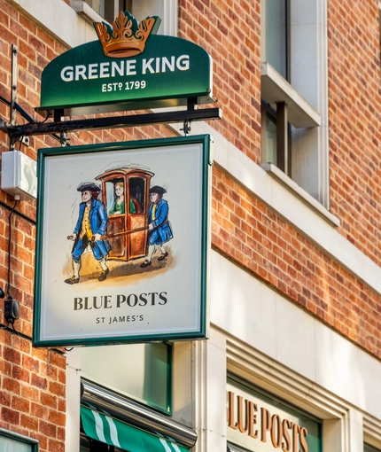 A close up view of the pub sign hanging on the exterior wall of the Blue Posts. The sign features the Greene King logo and an illustration of two people in blue coats carrying a woman in a sedan chair, above the words "Blue Posts, St James's". 