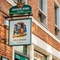 A close up view of the pub sign hanging on the exterior wall of the Blue Posts. The sign features the Greene King logo and an illustration of two people in blue coats carrying a woman in a sedan chair, above the words "Blue Posts, St James's". 