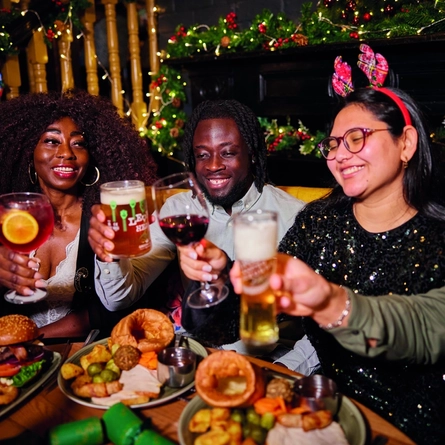 A group of people celebrating a festive Cheers while enjoying various main meals and drinks within the interior restaurant and seating area of Greene King Pub venue.
