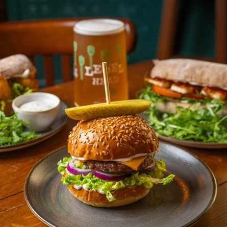 A Lunch Cheeseburger served on a plate which sits on a wooden restaurant table along with a glass of Level Head and other plates of food.
