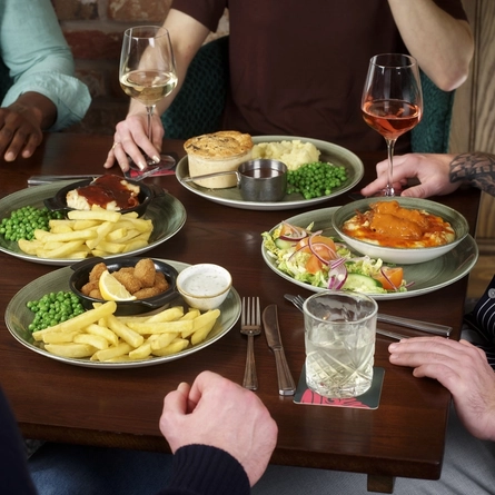 A round table filled with main dishes, including scampi and chips and a pie with mash and peas. Several drinks are placed around the table. The hands and arms of multiple people seated together are visible as they reach toward their food and raise their drinks.