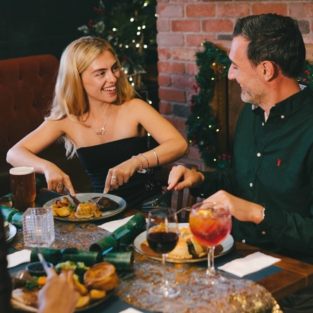 An image focusing on 2 people sat with friends enjoying various main dishes and a selection of drinks while sat within the interior restaurant seating area.