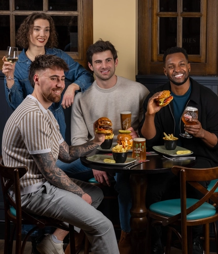 Four individuals sit around a restaurant table. Two individuals in the foreground each hold a large burger, with bowls of chips and condiments on the table. Another individual seated between them holds a tall glass of beer. A fourth individual standing behind the group holds a glass of white wine.