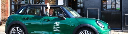 Larry Lamb waving his hand out of the driver's seat window of a green Mini Cooper, in front of a pub.
