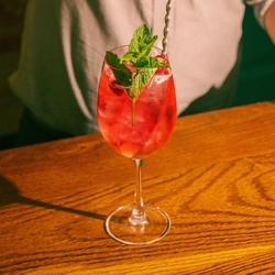 A Cherry Hugo Spritz cocktail served in a wine glass with ice, mint and dried raspberries; standing on the bar inside a pub. A bartender is stirring the cocktail with a long metal spoon.