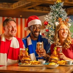 Three football fans sit around a wooden table in a sports pub decorated for Christmas, holding glasses of beer beside a frosted Christmas tree. One person wears a Santa hat and another wears reindeer antlers, while burgers topped with onion rings and pickle skewers, chips and dipping sauces are served on the table. Live football is shown on large screens in the background, creating a festive Boxing Day matchday atmosphere.
