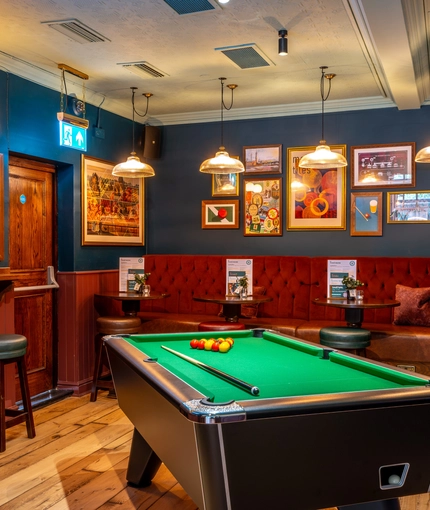 An interior restaurant seating and games area at the Phoenix in Bayswater; with a pool table, a TV on the wall, bar stool seating, and an upholstered booth seat along one wall.