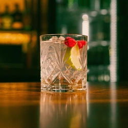 A close up view of a Coconut Margarita cocktail served in a whiskey glass with ice, dried raspberries, and a lime wedge. The glass stands on the bar inside a pub.