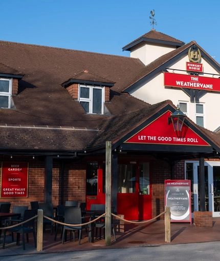 The exterior facade, signage, and seating area of The Weathervane, with wooden picnic benches, shade umbrellas, and string lights.