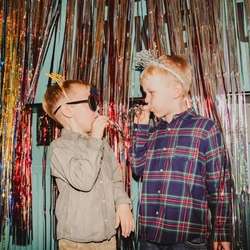 Two children stand in front of a shimmering gold and silver tinsel backdrop at a New Year’s Eve celebration, blowing party horns. One child wears a “Happy New Year” headband, while the other wears novelty New Year-themed glasses and a festive headband. The colourful metallic decorations and party accessories create a fun family-friendly New Year’s Eve scene.