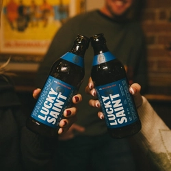 Two friends holding a bottle of low and no Lucky Saint beers in a cheers in a Greene King venue.