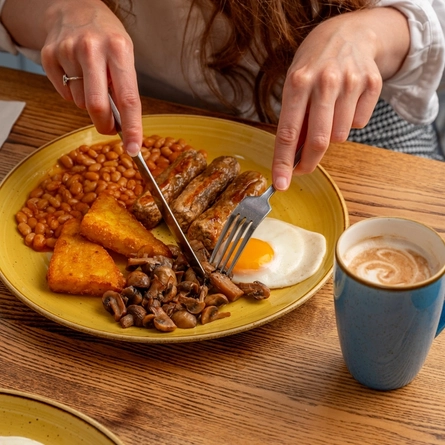 A shot taken above of a person's hands using cutlery over a plated cooked breakfast.