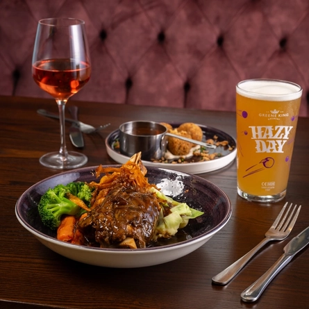 An advertising image of a bowled main and plated starter dish from the January/February Specials menu sat on a table with a selection of drinks within the interior restaurant seating area at a Community Pubs with Food venue.