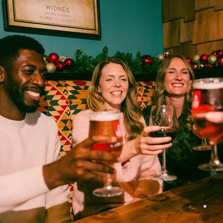 Four people sit together in a cosy pub booth decorated with Christmas garlands and red and gold baubles. The group raises glasses of lager and red wine in a toast over a wooden table, while festive drinks and seasonal décor create a warm and sociable Christmas pub atmosphere.