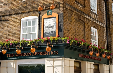 A close up view of the exterior facade and signage of The London Apprentice, with planter boxes filled with flowers above the golden yellow lettering spelling "The London Apprentice" on two sides of the pub.