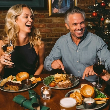 Three people sitting at a restaurant table with Christmas crackers and plates of food in front of them. One person is holding a glass of red wine, one person is holding a glass of white wine, and the third person is cutting up their food with their knife and fork.