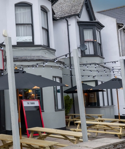 The exterior facade, signage, and seating area of The Tamar in Crownhill, with wooden picnic tables, shade umbrellas, and string lights overhead.