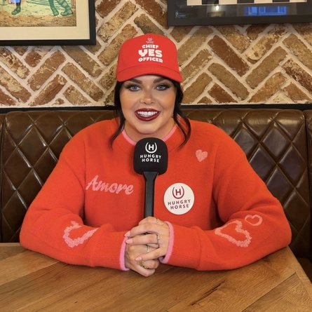 A portrait image of Scarlett Moffat sat in the interior dining area in a Hungry Horse venue wearing Hungry Horse branded long sleeve top and a cap with a Hungry Horse microphone held in both hands.