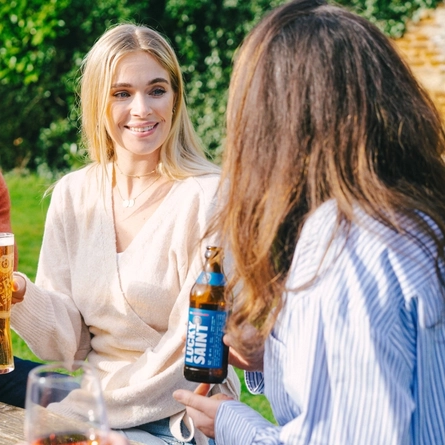Three people sitting at a wooden table in a sunny beer garden, one of them taking a sip from their glass of beer, one holding a glass of beer, and the other holding a bottle of Lucky Saint.