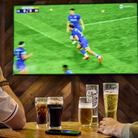 Two people sitting at a table in a pub, watching sports on the TV on the wall in front of them. A round of drinks and a mobile phone sit on the table between them.