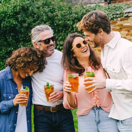 Four people standing in a beer garden, laughing together and each holding a glass of Pimm's cocktail garnished with mint leaves and fruit.