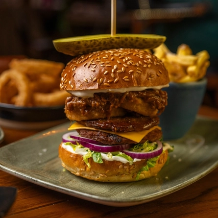 A close up view of a Pulled Pork Beef Burger served on a plate with a small bowl of fries. The plate sits on a wooden restaurant table and another plate of food is visible in the background.