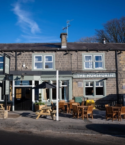The exterior facade, signage, and seating area of The Horsforth, with wooden picnic tables, shade umbrellas, and strings lights above the seating area.