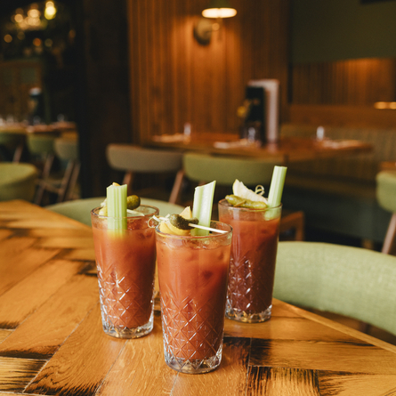 An advertising image showing 3 glasses containing Bloody Mary cocktails sat on a wooden table within the interior seating area at a Crafted venue.