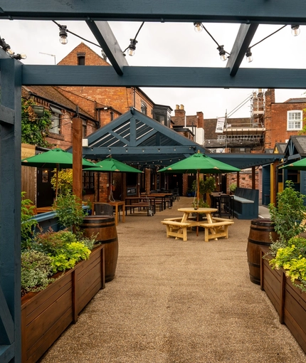 The exterior beer garden seating area at The Talbot; with wooden picnic tables, planter boxes, shade umbrellas, string lights overhead, and a large wooden pergola with under cover seating.