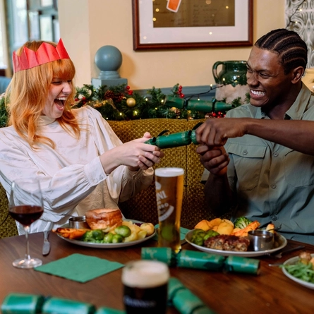 Two people seated at a table in a festively decorated pub pull a green Christmas cracker between them. One person wears a red paper crown and a light-coloured blouse, while the other wears a light green shirt. Plates of roast dinners, a pint of beer, a glass of red wine and additional Christmas crackers are arranged on the table. Seasonal garlands, fairy lights, framed artwork and patterned wallpaper create a festive dining setting.