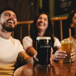 A pint of Guinness on the table in the interior seating area of a Crafted venue, three people are blurred in the background, one is holding the pint.