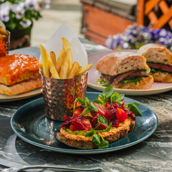 An advertising image showing three different sandwich dishes including Heritage Tomato Sandwich, Sirloin Steak Ciabatta, Panko Haddock & Ham & Emmenthal Croque Monsieur sat on a table within the exterior beer garden seating area at a Crafted venue.