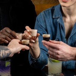 A group of people seated around an indoor table with a selection of drinks and shots in front of them.
